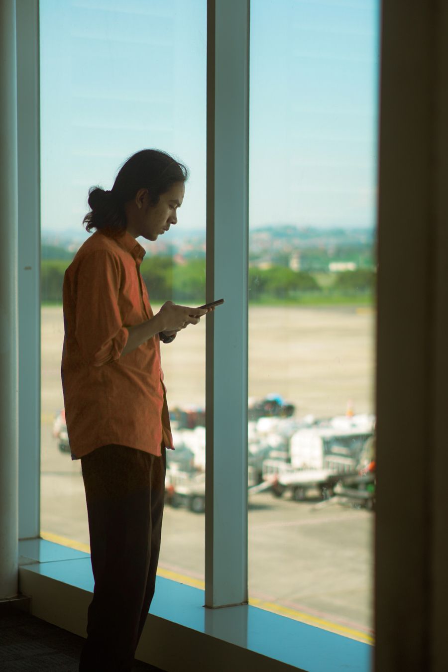 Man in orange shirt using a smartphone while standing at an airport window, overlooking the runway. — La escala en Venezuela: trabajar, streaming y mensajes, todo cubierto.