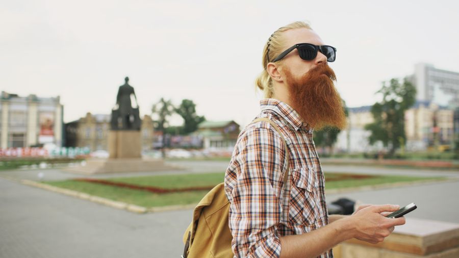 Hipster with a beard and sunglasses in a city square holding a smartphone. — El hall de llegadas de Chile: en línea antes de la cinta de equipaje, siempre.