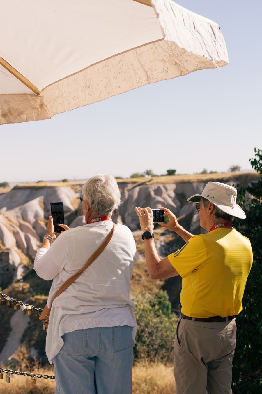 Senior tourists photographing scenic landscape in Cappadocia, Türkiye under a sun umbrella on a sunny day.