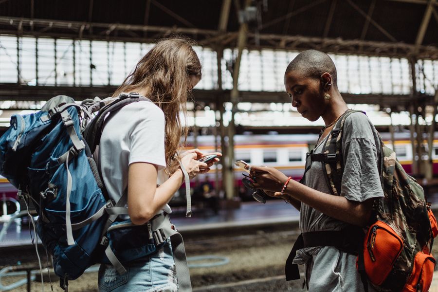 Two adventurous backpackers using smartphones while waiting at a bustling train station. — Caza de souvenirs en Panama con conversión de moneda en vivo en el bolsillo.